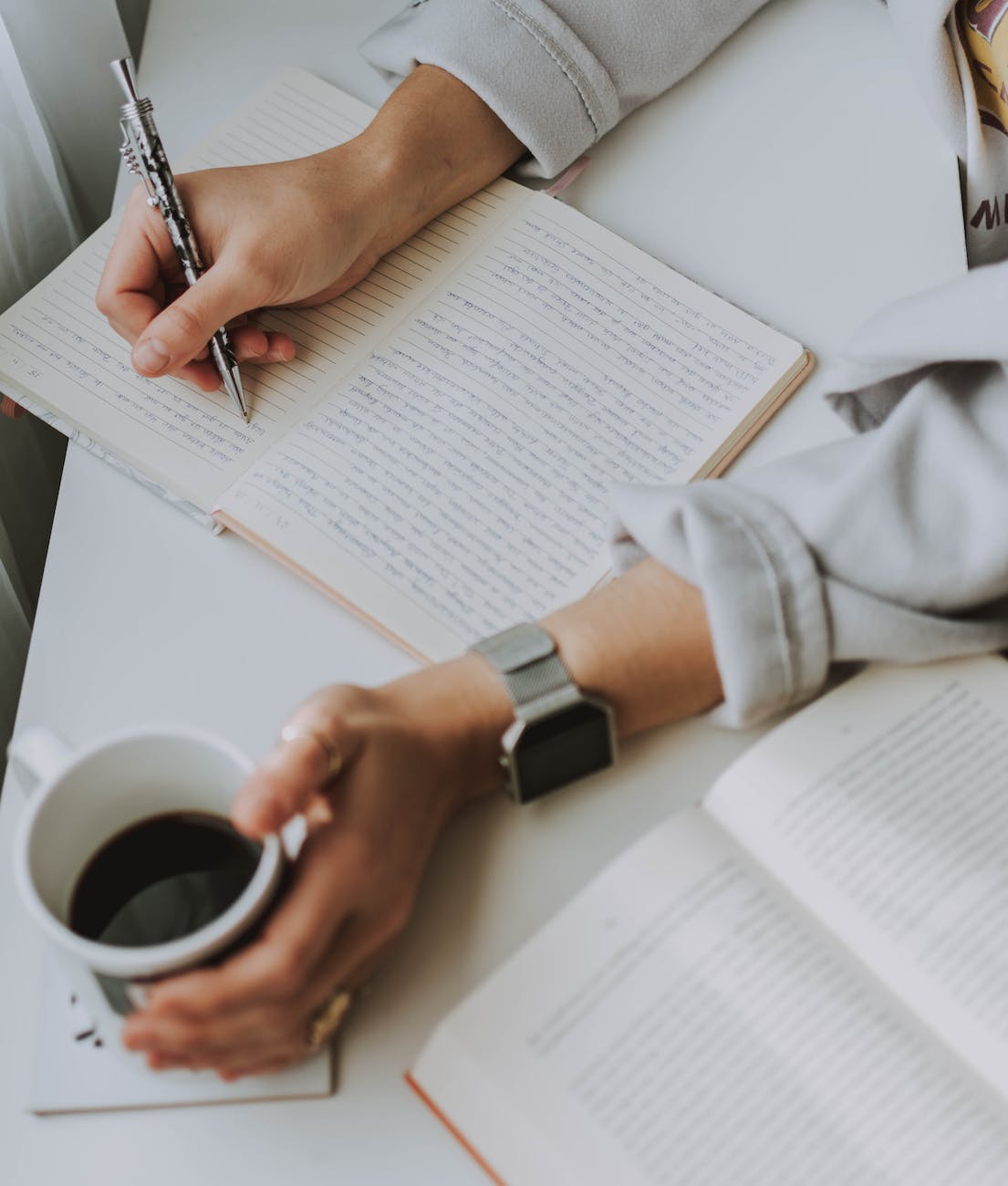 person writing on notebook while holding coffee mug